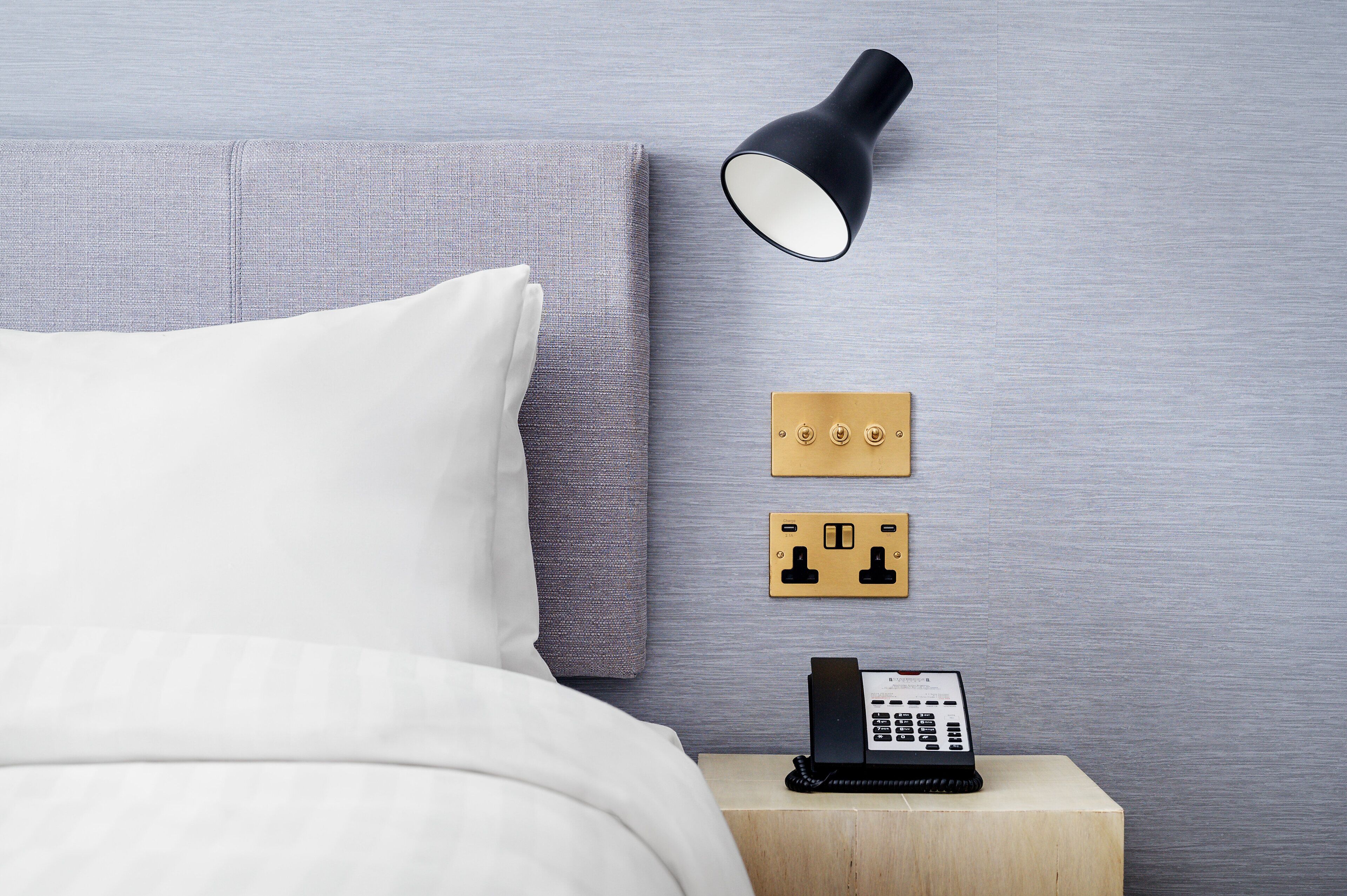 A neatly made bed with white linens, a bedside lamp, a wall-mounted telephone, and two electrical outlets on a wooden nightstand.