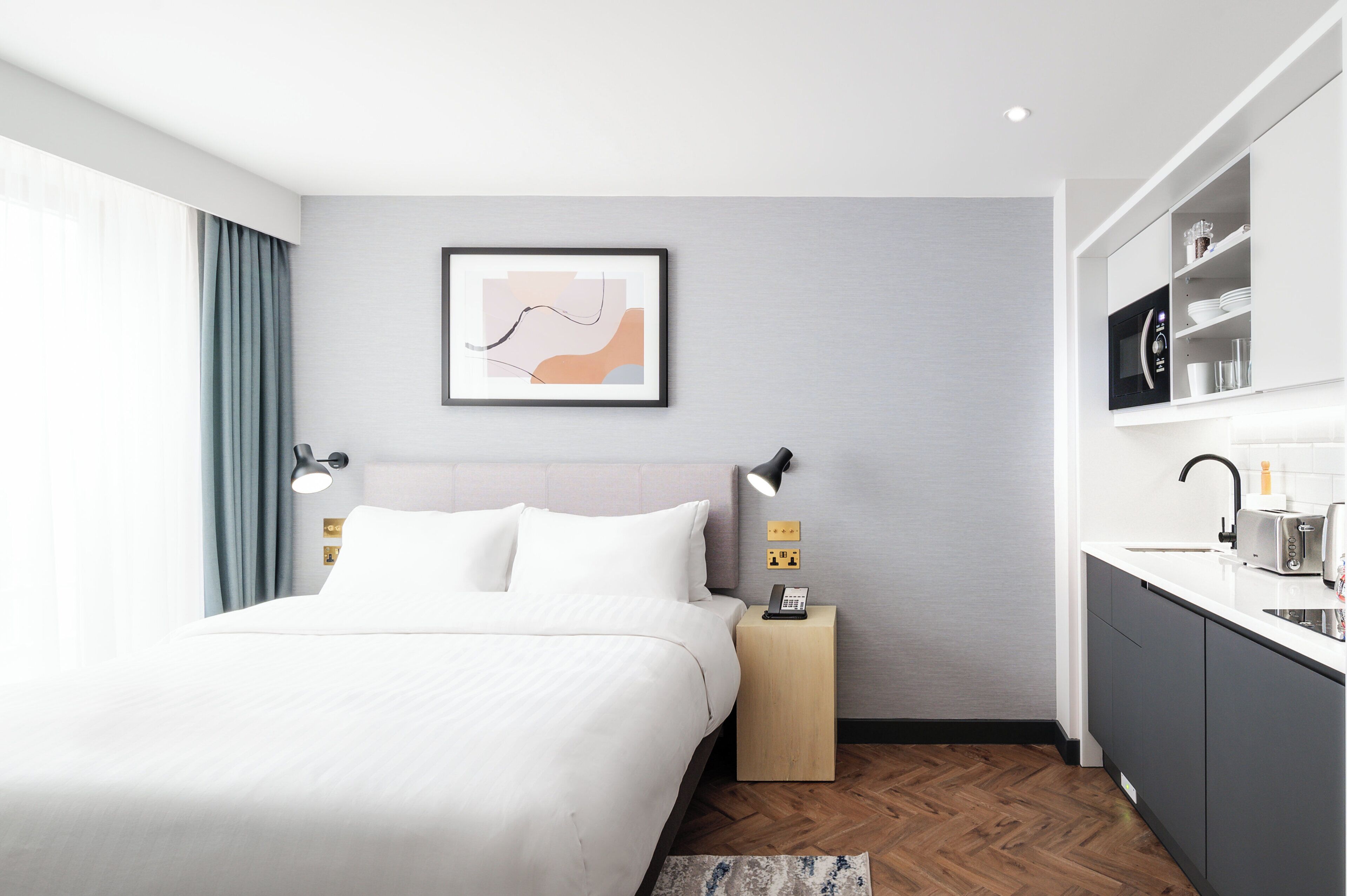 A modern hotel room with a bed, bedside lamps, a small bedside table with a phone, a framed abstract artwork on the wall, and a kitchenette area.