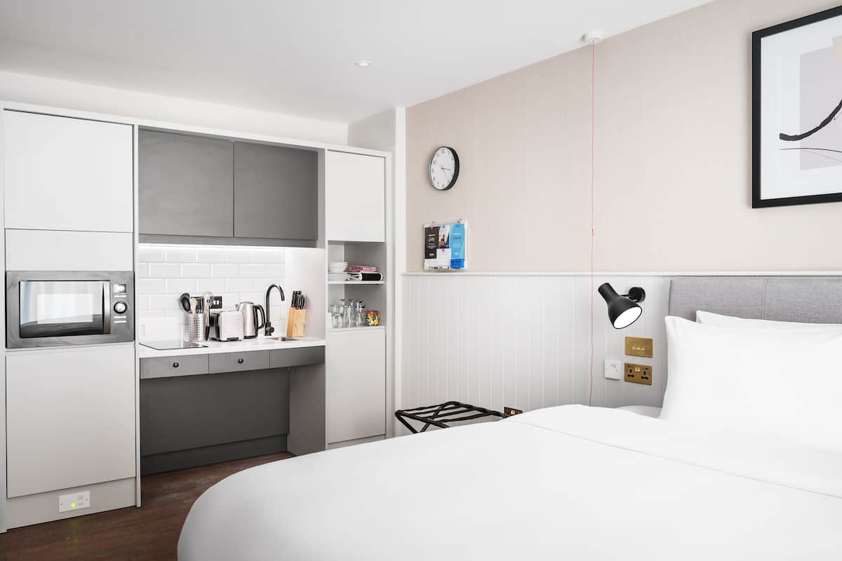 A modern hotel room with a kitchenette, a bed, a wall clock, and a framed picture.