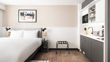 A hotel room with a bed, bedside table, a small stool, a kitchen area with a microwave, toaster, and kettle.