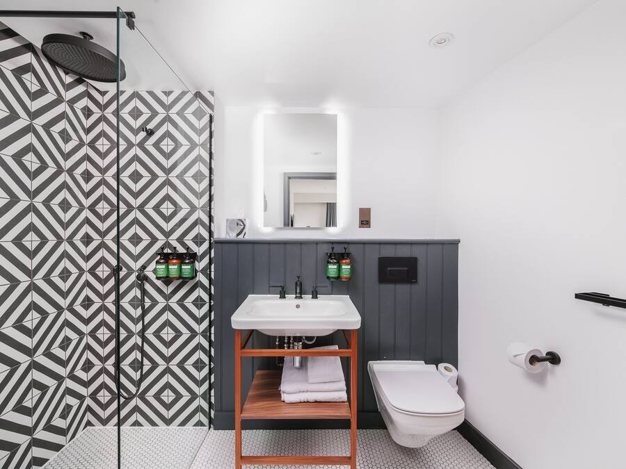 Ein modernes Badezimmer mit einer Dusche, deren Wand ein geometrisches Muster aufweist, einem weißen Waschbecken mit Holzablage und einem WC mit Handtuchhalter.