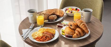 A breakfast table with croissants, toast, eggs, beans, sausages, and a fruit bowl.
