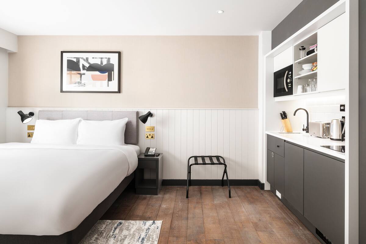 A modern hotel room with a bed, bedside table, nightstand, and a small stool.