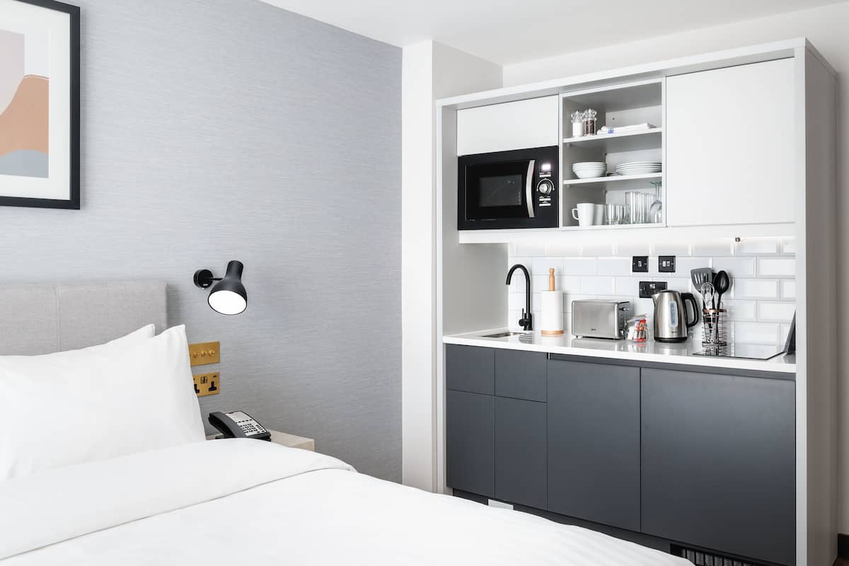A hotel room with a bed, a bedside table with a lamp, a phone, a microwave, a coffee maker, and a small kitchen area with cabinets and a sink.