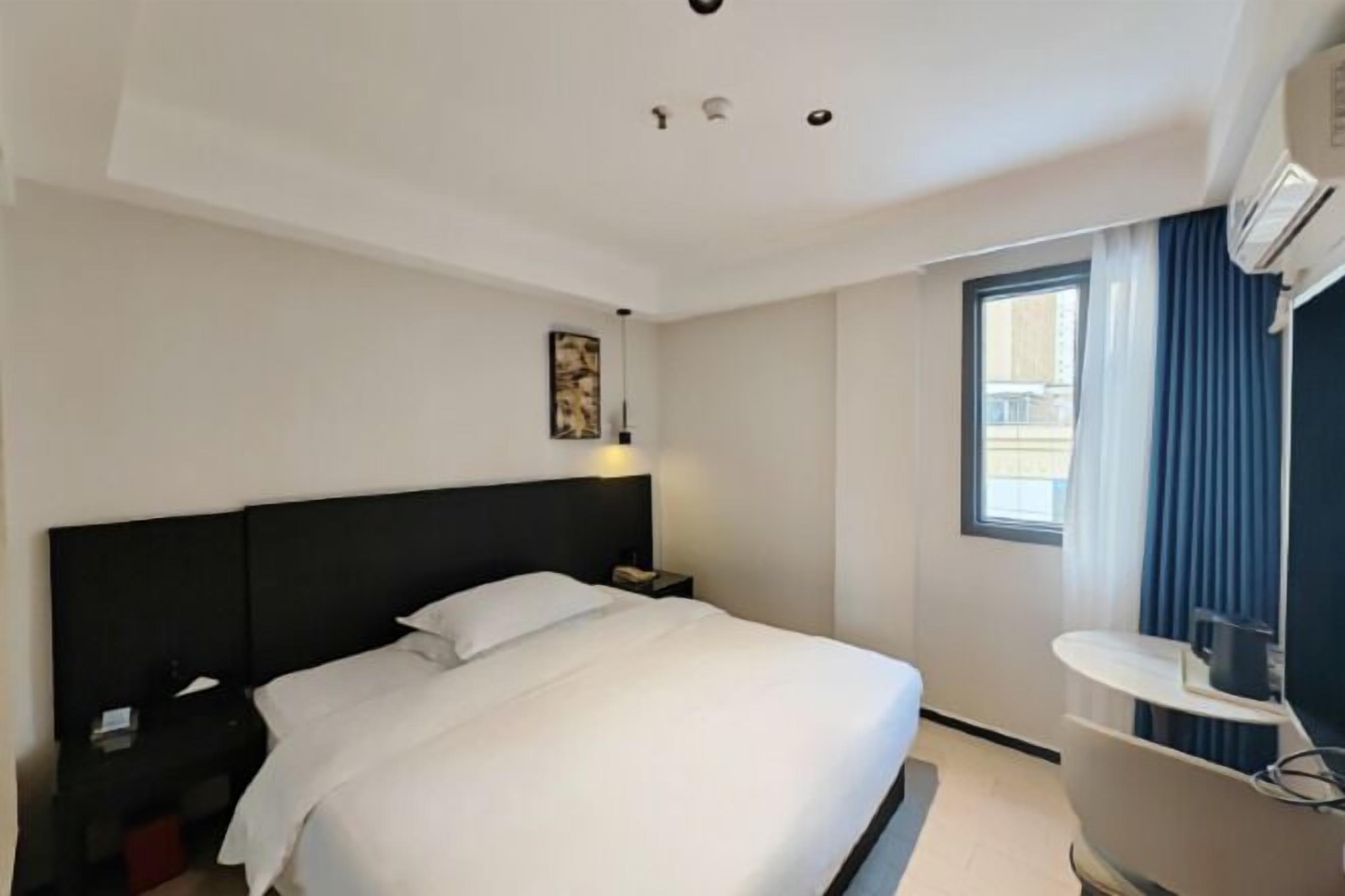 A hotel room with a bed, a nightstand, a desk, a window with curtains, and an air conditioning unit.