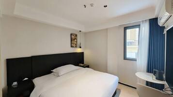 A hotel room with a bed, a nightstand, a desk, a window with curtains, and an air conditioning unit.