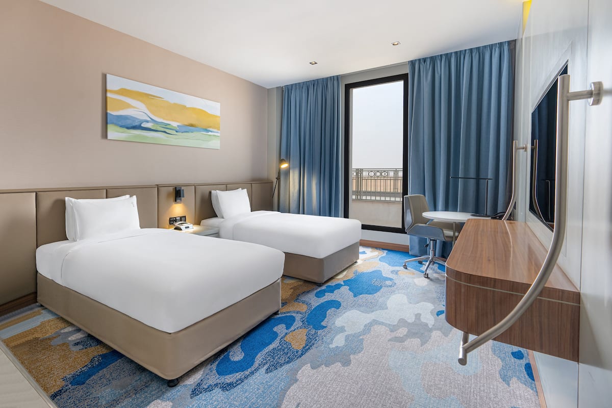A hotel room with two beds, a desk, a chair, a TV, and a balcony with curtains.