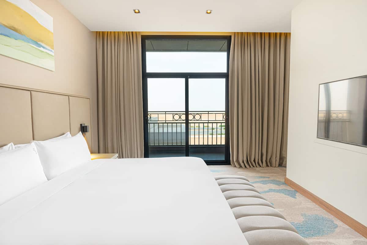 A hotel room with a large bed, a TV, a balcony with curtains, and a painting on the wall.