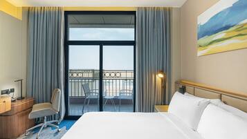 Habitación de hotel con una cama grande, un escritorio con silla, un balcón con sillas y un cuadro en la pared.