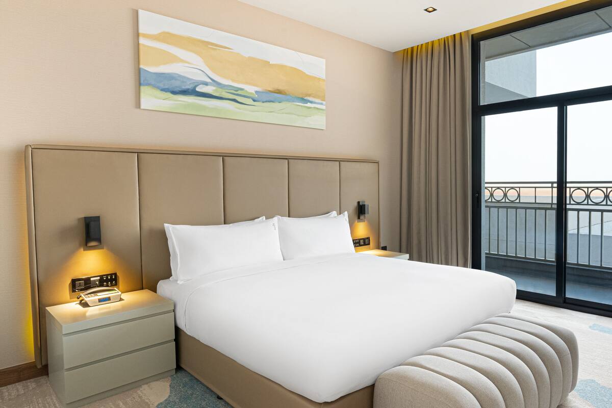 A hotel room with a large bed, bedside lamps, a nightstand, a painting on the wall, and a view through a large window.