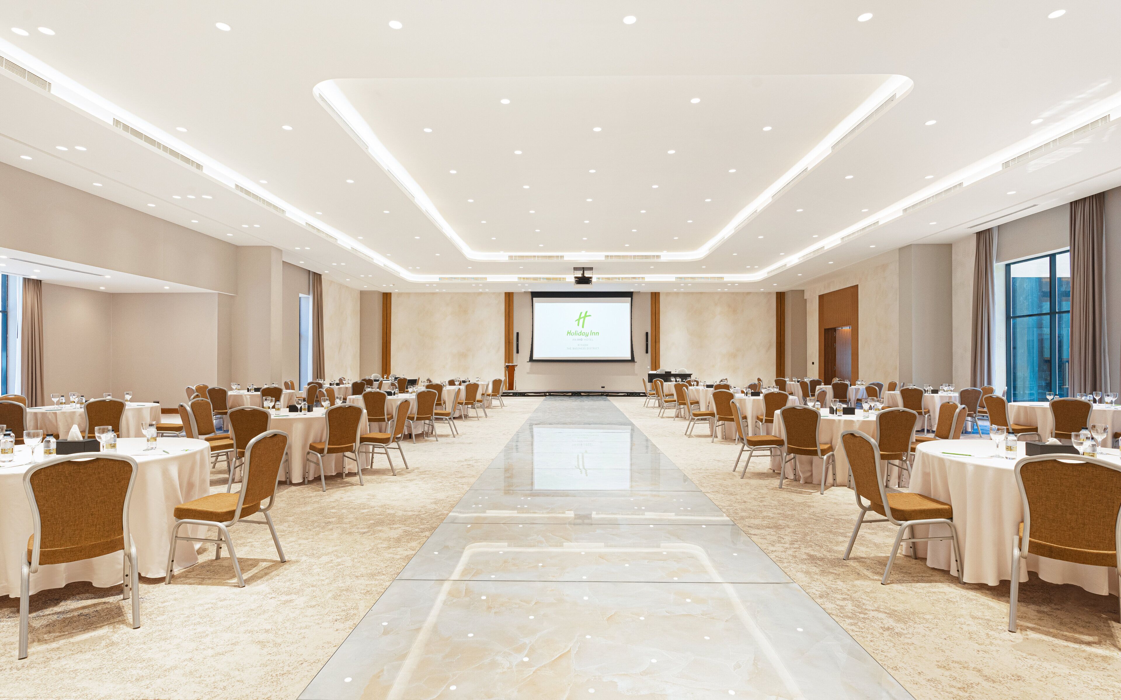 A conference room with a large screen displaying 'Holiday Inn' branding, arranged tables with chairs, and a polished marble floor.