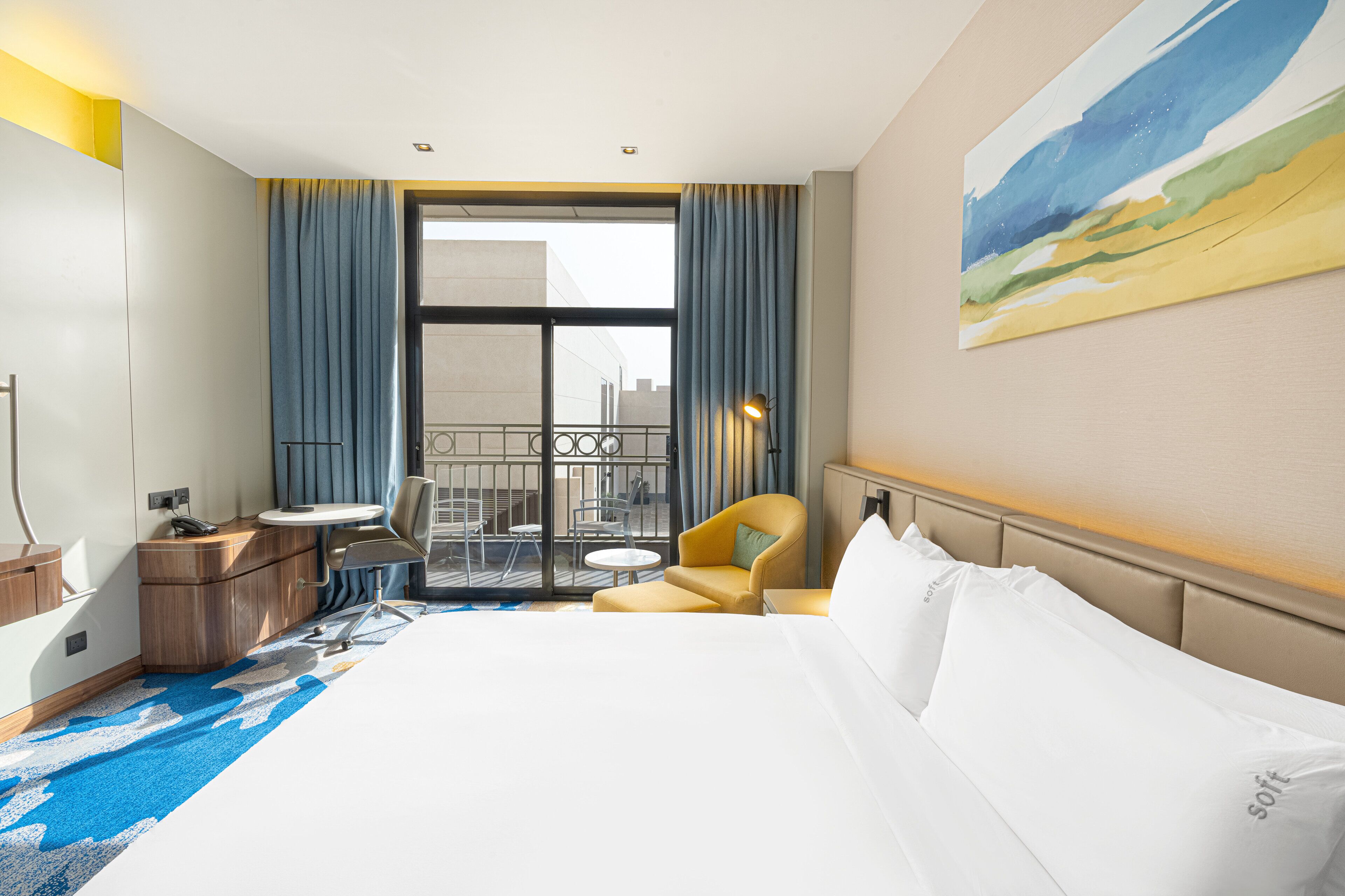 A hotel room with a large bed, a desk with a chair, a small table, and a balcony with a railing.