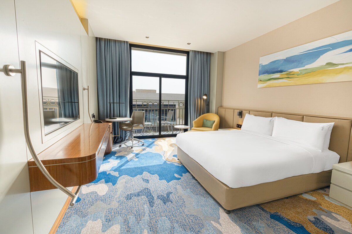 A hotel room with a large bed, a desk, a chair, a small table, and a balcony with a view.