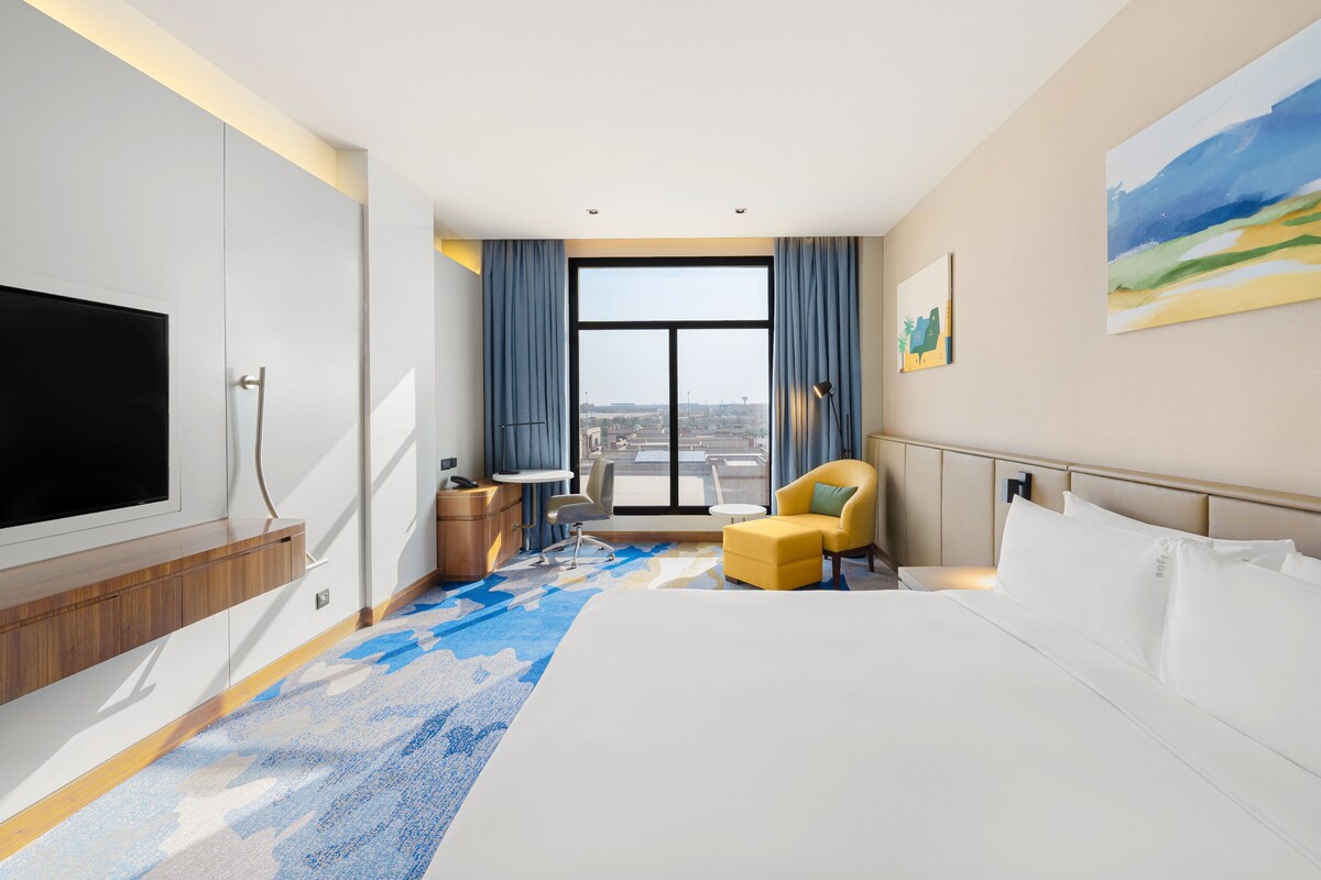 A modern hotel room with a large bed, a TV, a desk, and a view of the city through large windows.