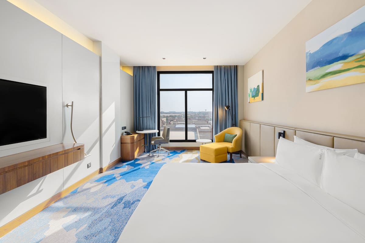 A modern hotel room with a large bed, a TV, a desk, and a view of the city through large windows.