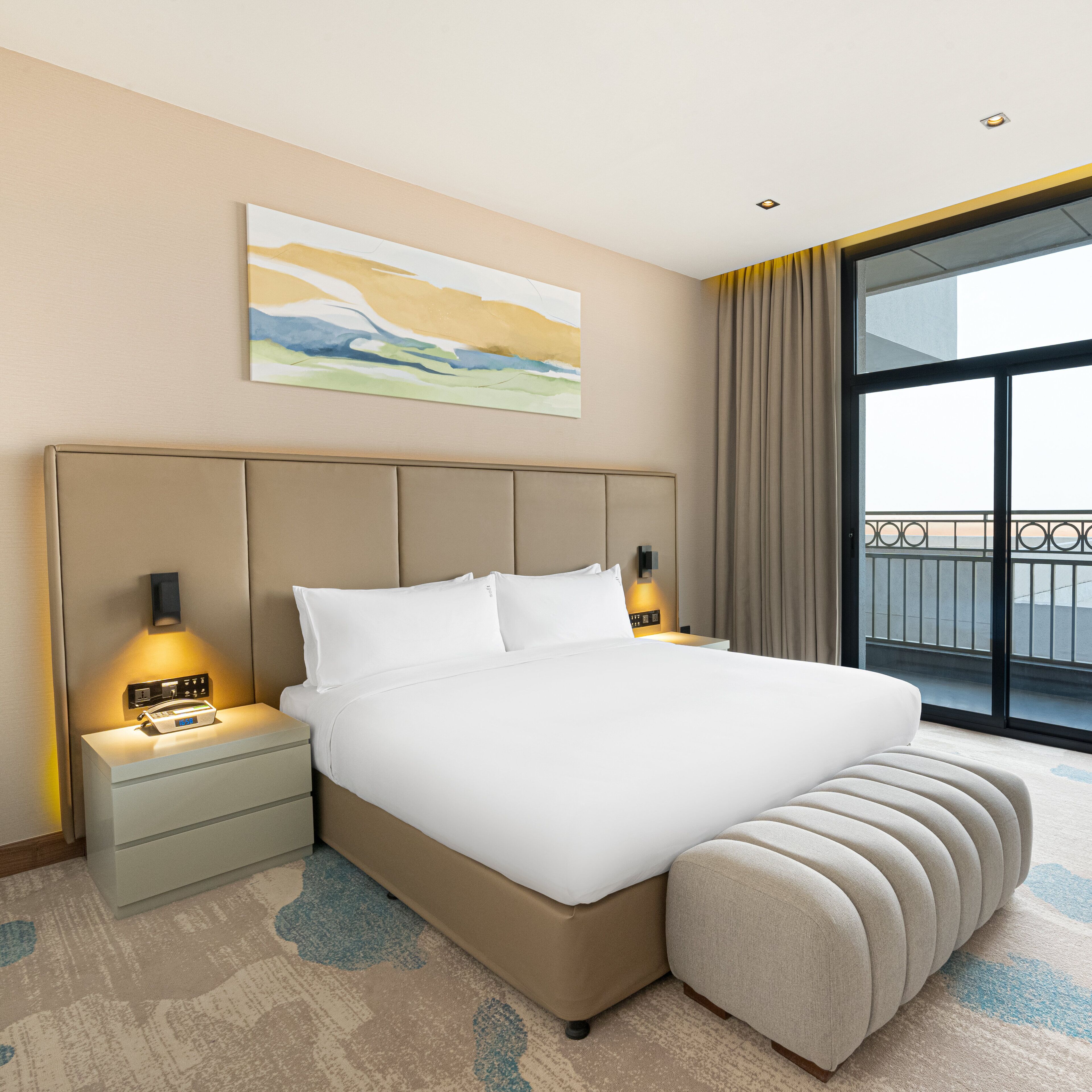 A hotel room with a large bed, bedside lamps, a nightstand, a painting on the wall, and a view through a large window.
