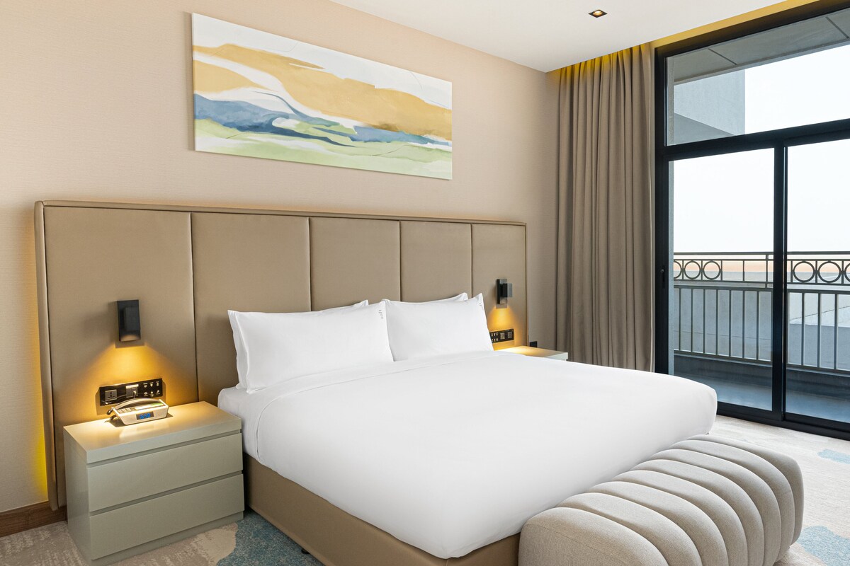 A hotel room with a large bed, bedside lamps, a nightstand, a painting on the wall, and a view through a large window.