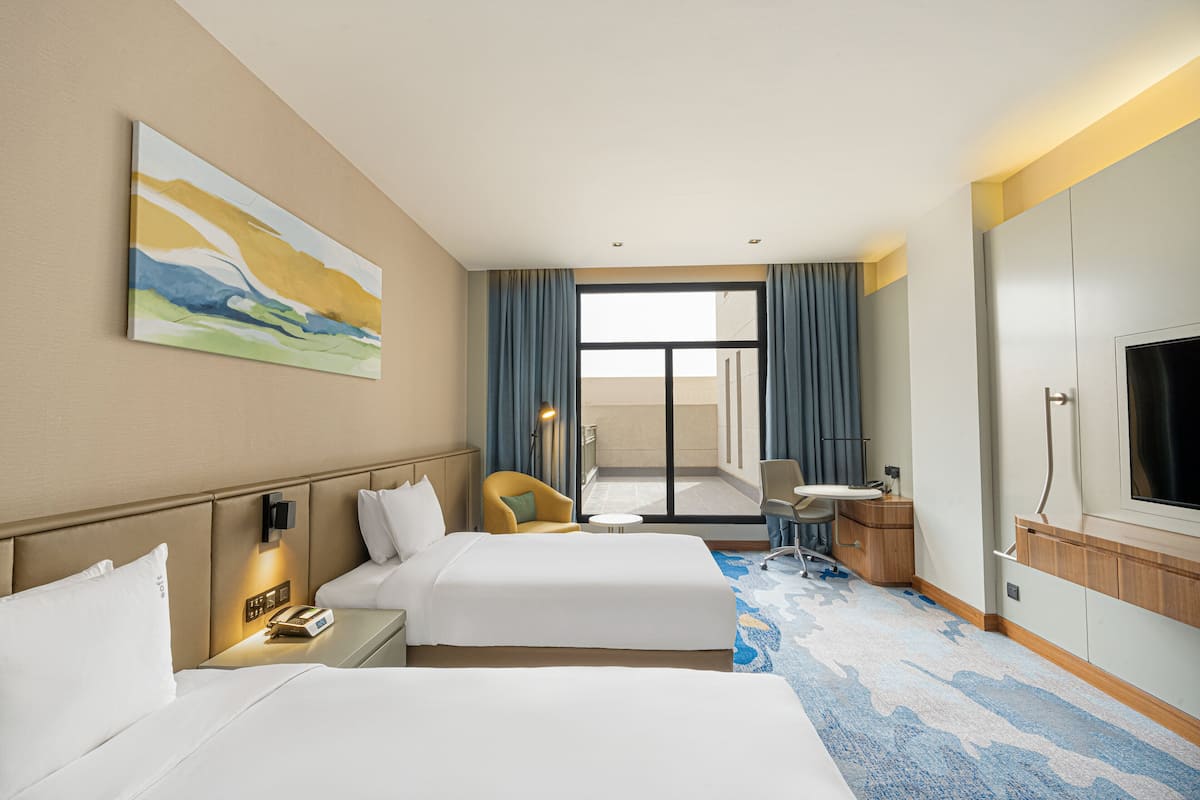 A hotel room with two beds, a desk, a chair, a TV, and a painting on the wall.