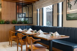Un restaurante con mesas y sillas de madera, un rincón con asientos tipo banca en tono azul oscuro, y ventanas amplias con vistas.