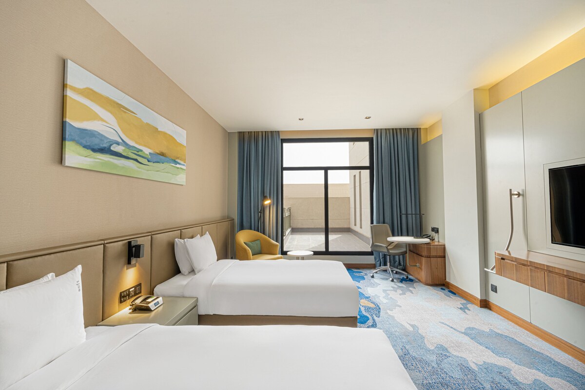 A hotel room with two beds, a desk, a chair, a TV, and a painting on the wall.
