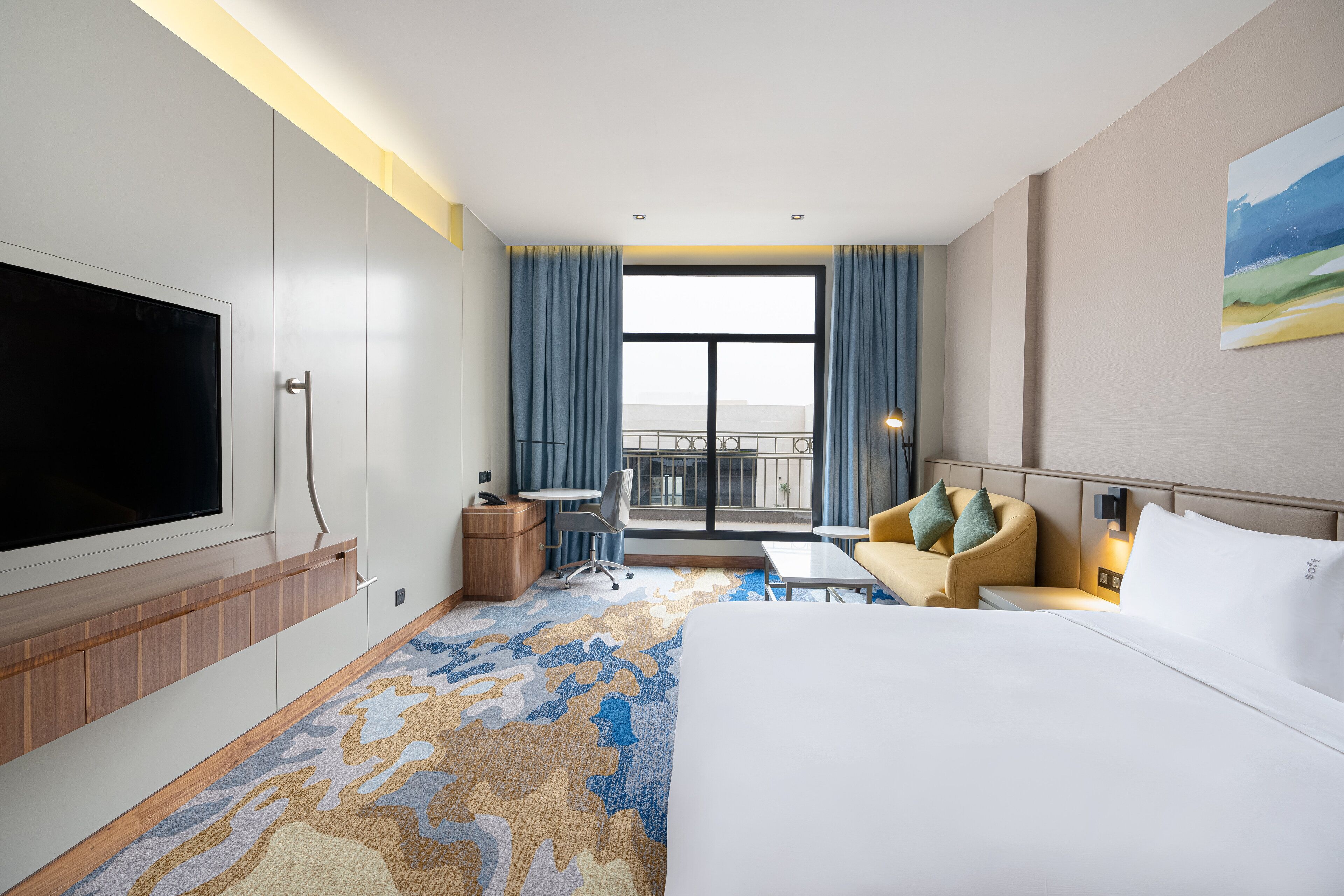 A hotel room with a large bed, a desk, a chair, a TV, and a balcony with a view.