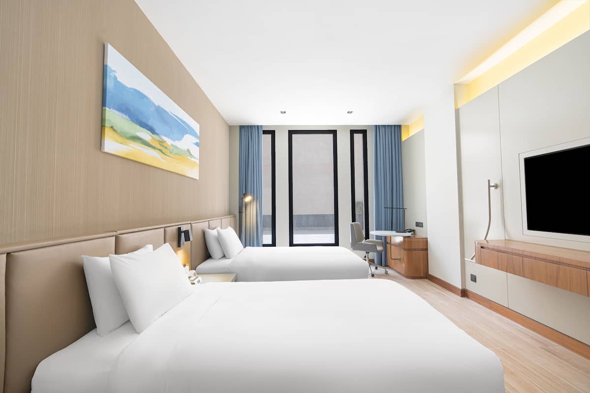 A hotel room with two beds, a desk, a TV, and a large window with curtains.