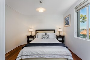 Une chambre à coucher bien aménagée, avec un grand lit, deux tables de chevet avec des lampes, une fenêtre donnant sur des immeubles et un luminaire au plafond.