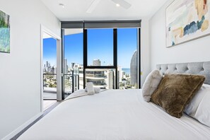 A modern bedroom with a large bed, a city view through floor-to-ceiling windows, and a ceiling fan. - Casino Private Apartments - QStay (Broadbeach)