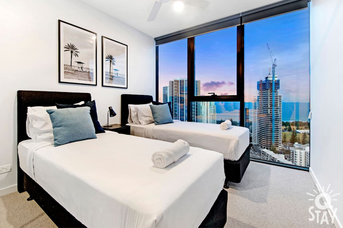A hotel room with two beds, a large window with a city view, and framed pictures on the wall.