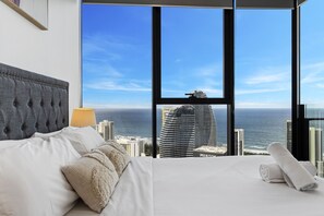 A hotel room with a large window, a bed, a lamp, and a view of the ocean and city skyline.