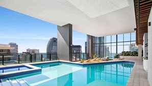 A rooftop infinity pool with clear blue water, surrounded by large windows offering city views. - Casino Private Apartments - QStay (Broadbeach)
