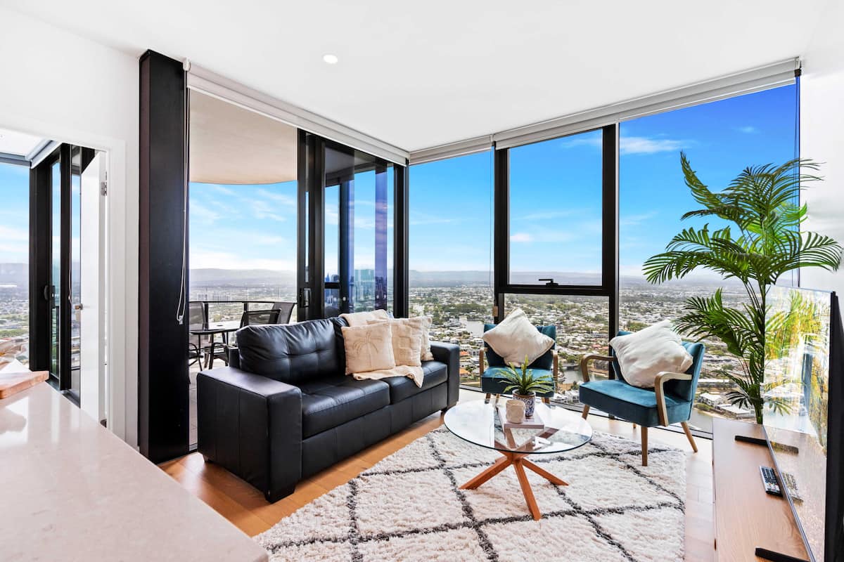 A modern living room with a black leather sofa, a round glass coffee table, and a view of the cityscape through large windows.