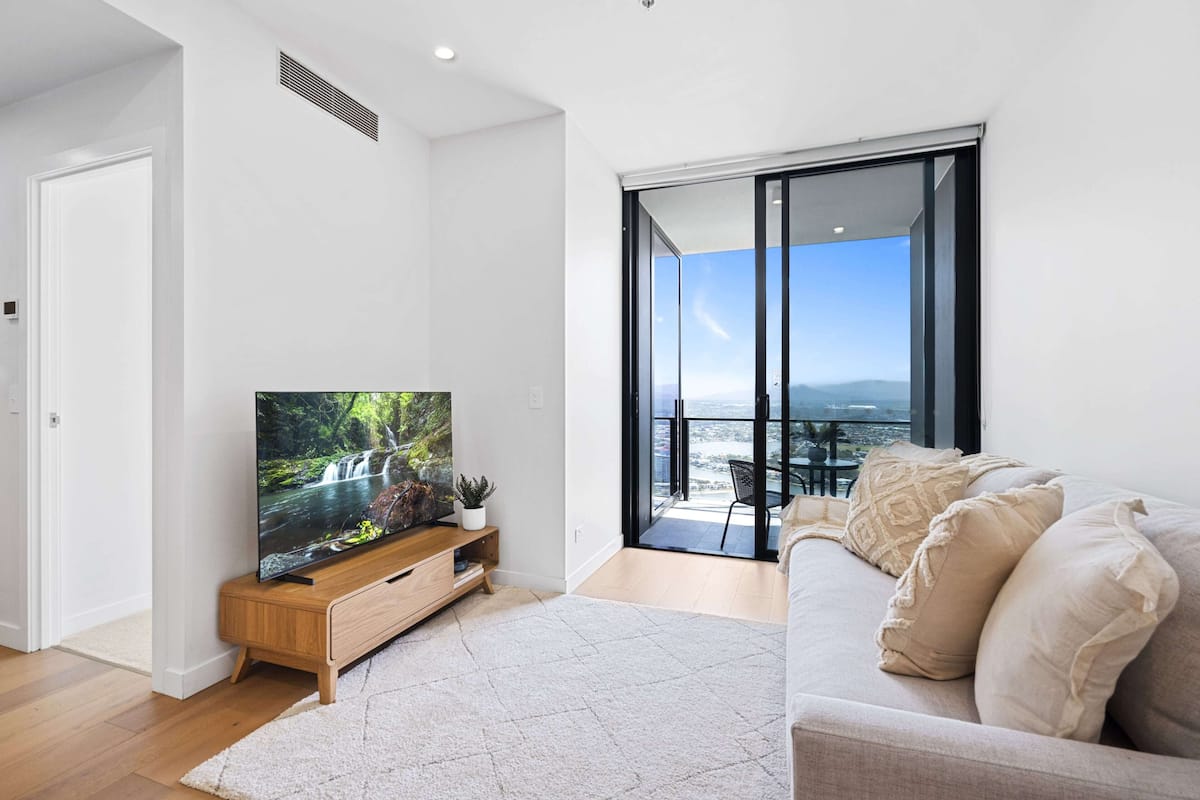 A modern living room with a flat-screen TV, a wooden entertainment unit, a beige sofa, and a large sliding glass door leading to a balcony with a view of the sea.