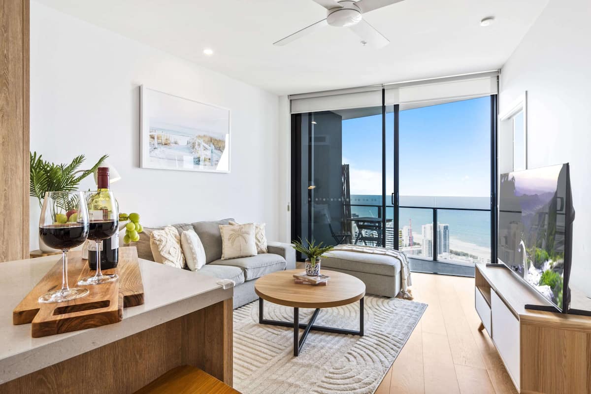 A modern living room with a sofa, a coffee table, and a TV. There is a balcony with a view of the city.