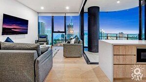 A modern living room with a city view, a large flat-screen TV, and a cozy seating area. - Casino Private Apartments - QStay (Broadbeach)