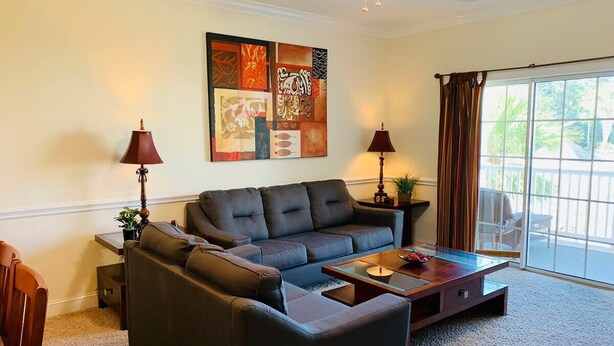 Ein Wohnzimmer mit einem dunkelgrauen Sofa, einem Glastisch und zwei Lampen. An der Wand hängt ein großes abstraktes Gemälde und eine Glastür führt in einen Außenbereich.