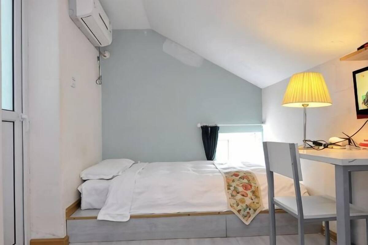 Une petite chambre d’hôtel composée d’une seule pièce, avec un lit, un bureau, une chaise et une lampe.