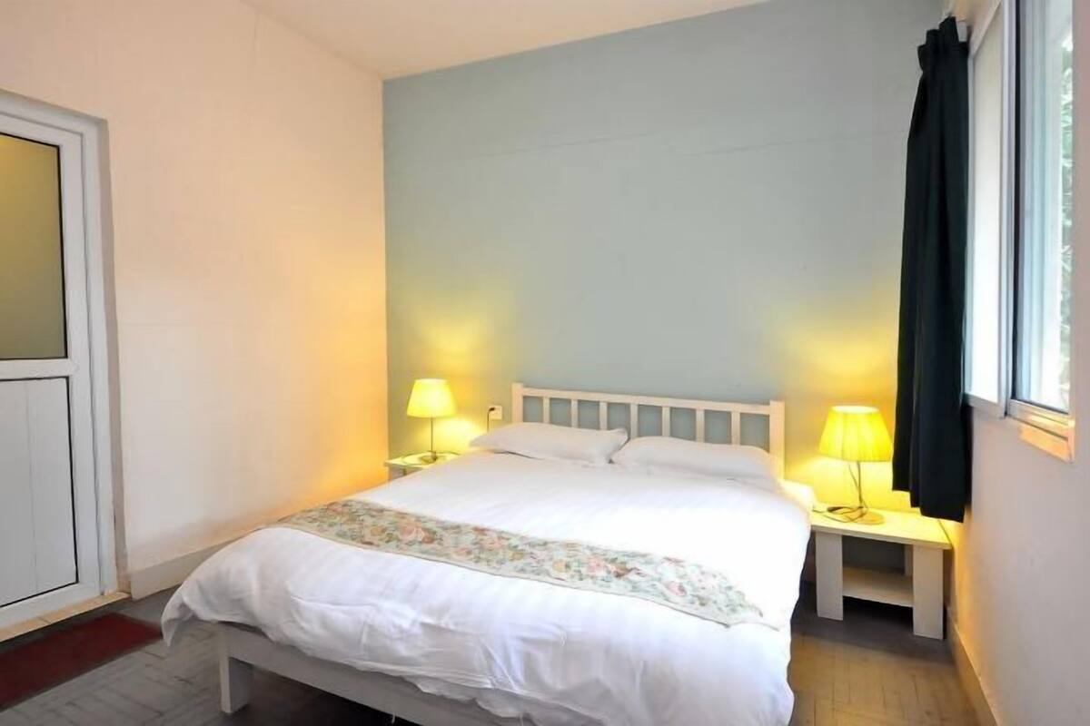 Un lit simple avec une tête de lit blanche, deux tables de chevet avec des lampes, une couvre-lit à motifs floraux et une fenêtre avec des rideaux noirs.