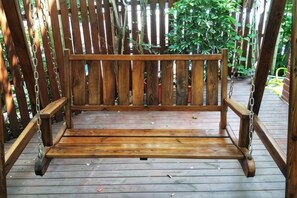 Een houten bank met een metalen ketting om iets aan op te hangen, op een houten terras met een houten hek op de achtergrond.