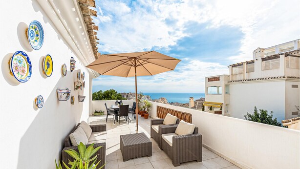 Un balcón con mobiliario de mimbre, una mesa, sillas y una sombrilla. Se observan platos decorativos en la pared y vistas al mar.