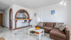 Una sala moderna con un sofá marrón, una mesa de centro blanca con un tazón de naranjas, y una zona de cocina con armarios blancos y un arco de ladrillo.