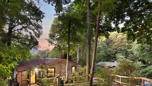 A rustic cabin with a stone exterior, surrounded by dense forest. A wooden deck leads to the entrance, and a small bridge crosses a gravel path. - High-Speed WiFi, Pool Table, private HOT TUB, Fire Pit, Game Room, 2 Kitchens! (Maggie Valley)