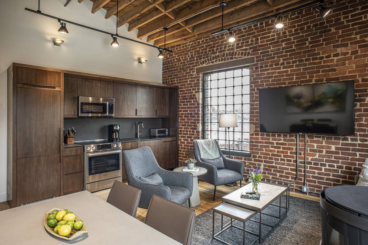 A modern kitchen with stainless steel appliances, a dining area with a table and chairs, and a brick wall with a mounted TV.