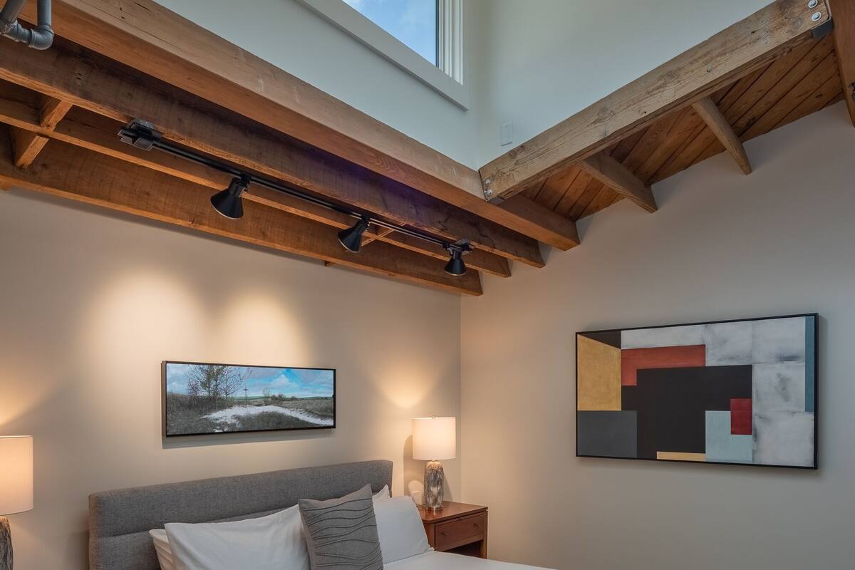 A bedroom with a large bed, a skylight, a painting, and a modern abstract art piece.