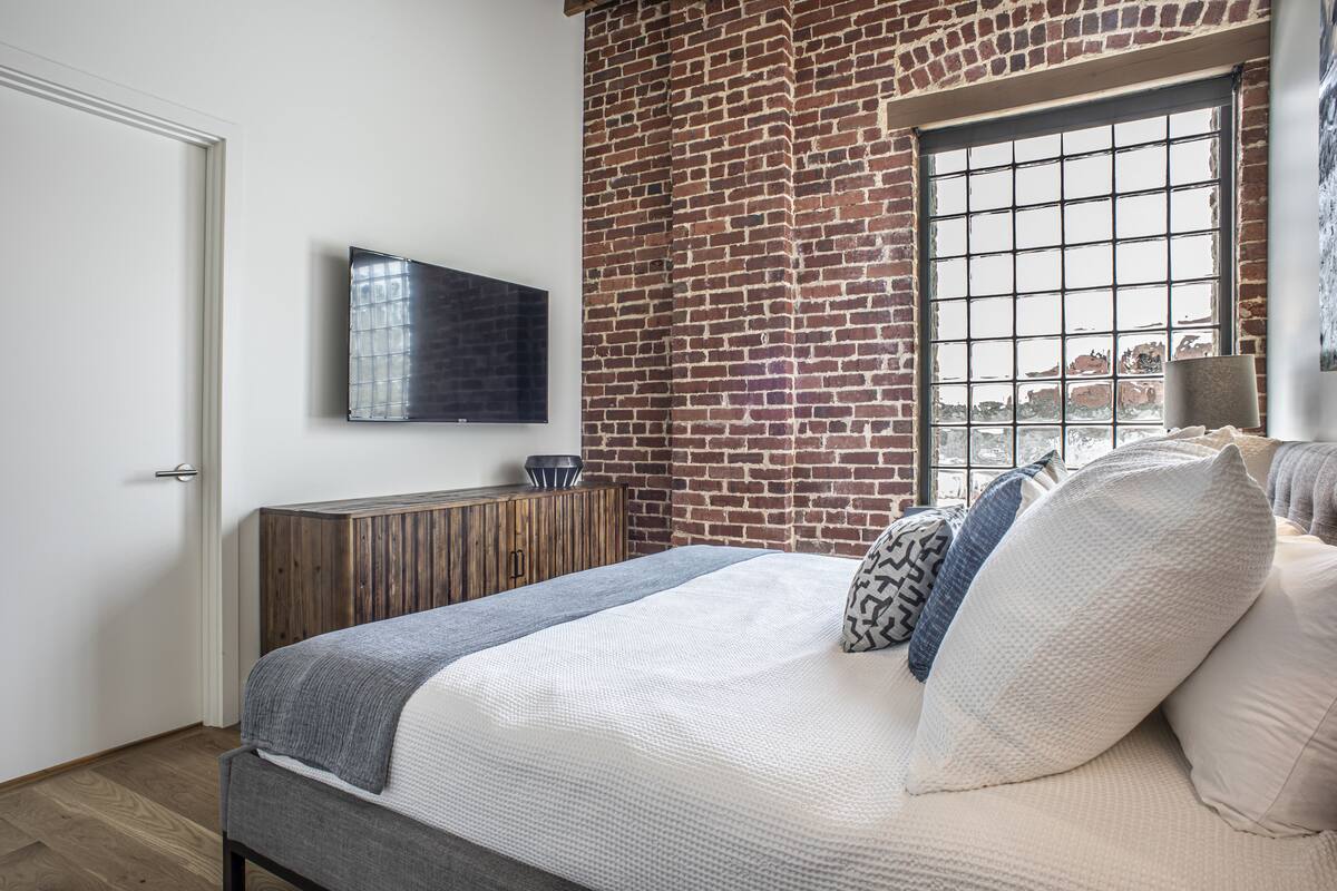 A bedroom with a brick wall, a bed with multiple pillows, a wooden cabinet, and a TV mounted on the wall.