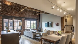 A modern living space with a brick wall, large windows, a fireplace, and a dining area. - Elevation Lofts Hotel (Asheville)