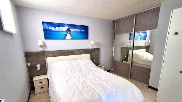 Quarto de hotel com cama, mesas de cabeceira, um quadro de paisagem marítima, guarda-roupa e espelho.