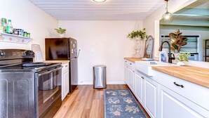 A compact kitchen with a stove, oven, refrigerator, and white cabinets. There is a trash bin and a wooden countertop with a sink.