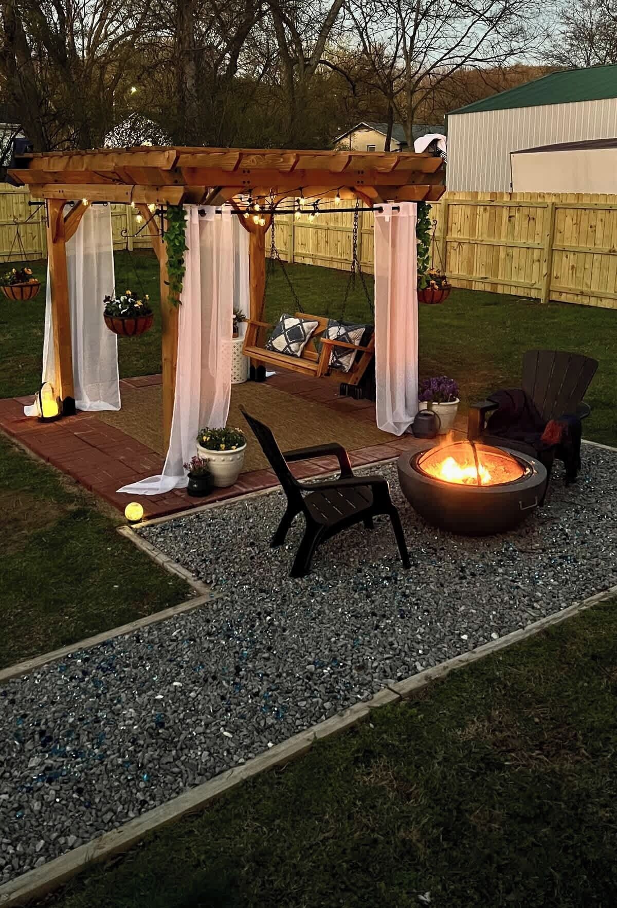 A patio with a fire pit, chairs, and a pergola with draped fabric.