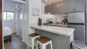 A compact kitchen with grey cabinets, a stainless steel refrigerator, a countertop with a kettle, and two stools.